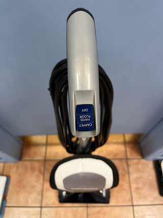 Top-down view of a Sew & Vac Oreck Elevate Conquer certified refurbished vacuum cleaner with its handle facing upwards, cord wrapped around, and a digital display reading "charge full" upside down.