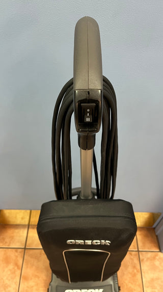 An upright Sew & Vac Oreck XL21 vacuum cleaner with a neatly coiled hose and a HEPA CC bag attached to its handle, against a blue wall.