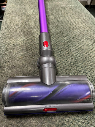 A close-up of a certified refurbished Sew & Vac Dyson Cyclone v10 Animal vacuum cleaner head on a carpeted floor, featuring a red button and clear dust container.