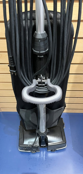 Certified reconditioned Sew & Vac Oreck XL21 upright vacuum cleaner with multiple cords neatly wrapped around its holder, placed on a blue surface against a wooden panel background, perfect for everyday cleaning.