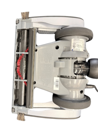 Bottom view of the refurbished Oreck Magnesium LW1500 vacuum head by Imagine Sewing & Vacuum, featuring wheels, a new red-bristled brushroll, and labels on a light grey plastic casing.