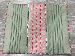 A rectangular quilted fabric placemat featuring vertical panels of green and pink patterns, bordered with a red scalloped trim, created using the precision of the Sew & Vac Baby Lock Applaud Creative Coverstitch & Serger Extravaganza machine.