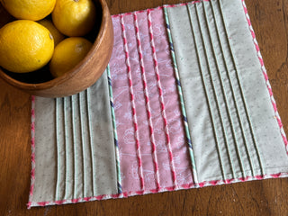 A wooden bowl filled with lemons sits on a wooden table next to a quilted placemat featuring pink and gray fabric sections, meticulously stitched using the Sew & Vac Baby Lock Applaud Creative Coverstitch & Serger Extravaganza.