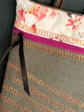 A close-up of a fabric pouch with a floral-patterned upper trim, crafted using the Sew & Vac Baby Lock Applaud Creative Coverstitch & Serger Extravaganza. It features a purple zipper and a black ribbon pull. The main body has a striped design with textured patterns, showcasing the precision of the coverstitch machine used in its creation.