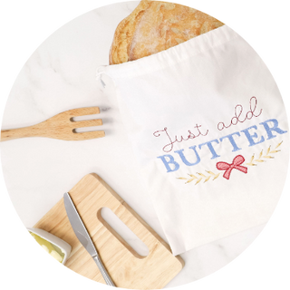 A cloth bag labeled Kimberbell "Just Add Butter" from the Layton In-Store Class 1-Day Event (February 28, 2026) holds a loaf of bread beside a wooden fork, cutting board, and butter knife on a white surface, evoking cozy cottagecore vibes.