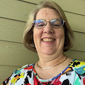 A woman with short, light brown hair and glasses smiles while wearing a colorful, floral-patterned shirt. She stands in front of a beige wooden wall, showcasing her latest creation—the Marrakesh Pillow—made using the Husqvarna Viking Epic 3 Embroidery Machine Sales Event from Sew & Vac.