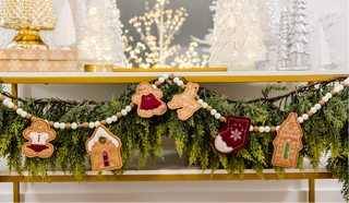 The Kimberbell Gingerbread Christmas garland by Sew & Vac features cookie-shaped gingerbread ornaments—including a house, gingerbread person, stocking, and tree—strung along a mantle adorned with greenery and bead accents to create the perfect Gingerbread Christmas atmosphere.