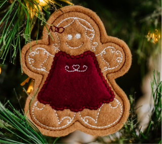 A felt gingerbread woman ornament with white stitching and a red dress hangs on a Christmas tree branch, adding a touch of charm to your Kimberbell Gingerbread Christmas-themed decorations by Sew & Vac.