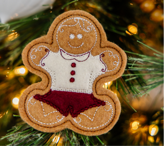 A felt gingerbread person ornament from Sew & Vac's Kimberbell Gingerbread Christmas collection, dressed in a white shirt with red buttons and maroon shorts, hangs on a Christmas tree adorned with white lights, adding charm to your holiday décor.