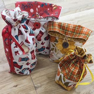 Four fabric drawstring bags from The Giving Bag Class Nov 13 2026 2-4 pm, featuring seasonal prints with autumn leaves, plaids, and Christmas stockings, secured with buttons or ribbon ties.