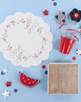 Create a festive look with the Kimberbell Patriotic Parade One-Day Event by Layton In-Store Class, featuring a white Redwork Table Round with red rocket and floral designs, fabric flowers in a red pail, wooden box, felt watermelon, and star decor.