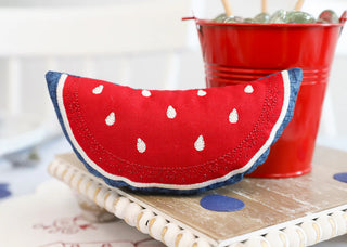 A watermelon slice fabric decoration sits beside a red bucket of marbles on wood—a fun accent for your Redwork Table Round or centerpiece at the Kimberbell Patriotic Parade One-Day Event by Layton In-Store Class, May 9, 2026.