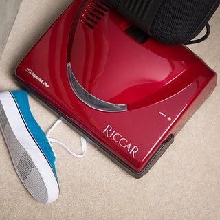 A Riccar R10P Lightweight Supralite Premium upright vacuum cleaner in red rests on the carpet, partially covering a blue and white sneaker.