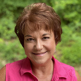 A person with short brown hair smiles, wearing a pink top that seems inspired by the intricate designs of the Viking Husqvarna Treasure Tote Embroidery Event from Sew & Vac. Greenery in the blurred background suggests an outdoor setting.