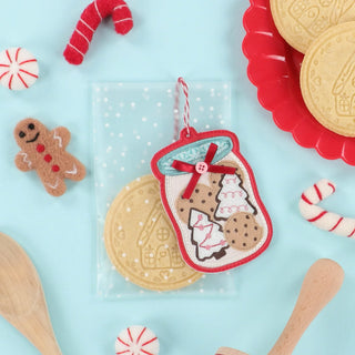 A felt ornament shaped like a jar of cookies is displayed alongside sugar cookies, peppermint candies, a felt gingerbread figure, and wooden utensils on a light blue background—offering perfect inspiration for your next creative session at the Kimberbell Mrs. Claus’ Bakery One-Day Layton Event, hosted by Layton In-Store Class on October 10, 2026.