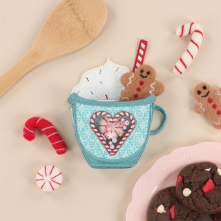 A delightful blue cup topped with whipped cream, gingerbread men, and a straw is surrounded by cookies, felt candy canes, a mint candy, and a wooden spoon on a beige surface—capturing the festive spirit of the Kimberbell Mrs. Claus’ Bakery One-Day Layton Event on October 10, 2026. This charming scene is perfect inspiration for your next holiday embroidery project during the Layton In-Store Class.