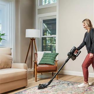 A person vacuums a living room rug with the Riccar R65 Cordless Stick Vacuum, featuring SupraCyclonic™ filtration. The room has a beige sofa, a wooden chair with cushions, a floor lamp, and large windows.