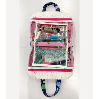 A clear zippered pouch with white and pink trim, created in the Layton In-Store Truvy Bag Class with Carla on 10/25/25, neatly holds pens, markers, a stapler, binder clips, and more—perfect for organizing sewing or stationery essentials.