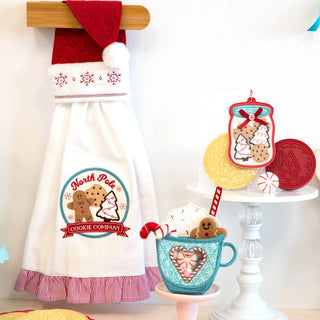 A white towel embroidered with “North Pole Cookie Company” hangs on a wooden rod, accompanied by a blue cup filled with felt cookies, an ornament, and red cookies arranged on a tiered stand—perfect inspiration for your holiday embroidery projects at the Kimberbell Mrs. Claus’ Bakery One-Day Layton Event on October 10, 2026 from 1000-Education.